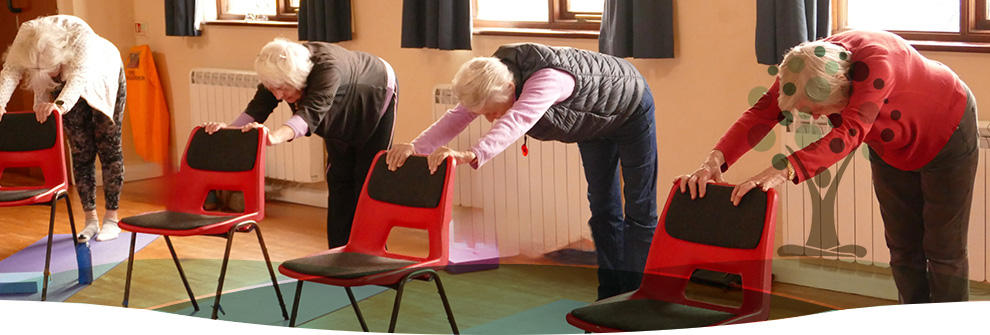 A chair-based class: four students holding the backs of red hall chairs and folding forward
