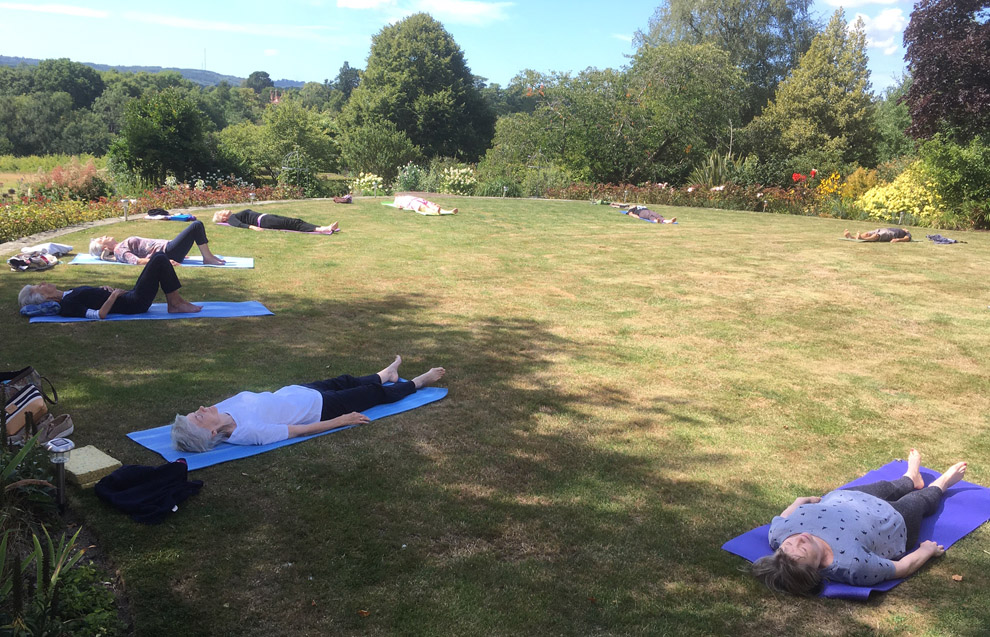 An outdoor yoga class on a lawn: students lying on blue and purple mats with a hedgerow and the Weald behind